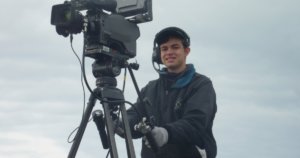 Ben Rosloff wearing a jacket and a headset standing behind a large video camera