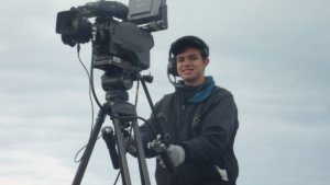 Ben Rosloff wearing a jacket and a headset standing behind a large video camera