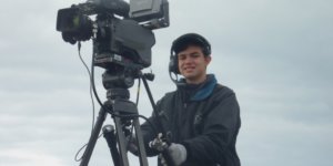 Ben Rosloff wearing a jacket and a headset standing behind a large video camera