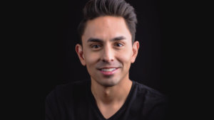 Danny Gomez smiling headshot wearing a black shirt in front of a black backdrop