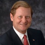 Steve Bartlett, has brown hair and he is smiling and wearing a black suit, white shirt, and red spotted tie and an american flag pin on his jacket, color photo