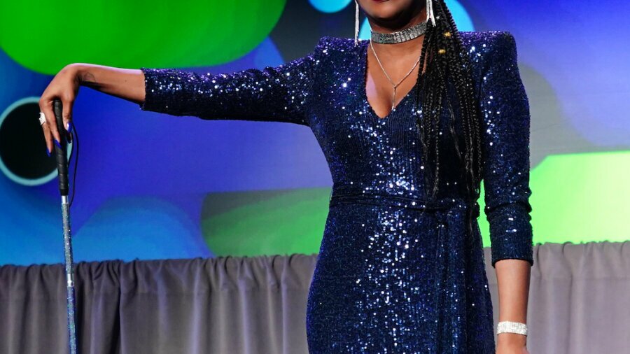 Lachi speaking on stage at an event wearing a sparkling blue dress and holding her cane