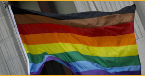 an LGBTQ+ Pride flag with black, brown, red, orange, yellow, green, blue and purple stripes