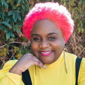 Nikki Bailey headshot smiling with pink hair wearing a yellow shirt