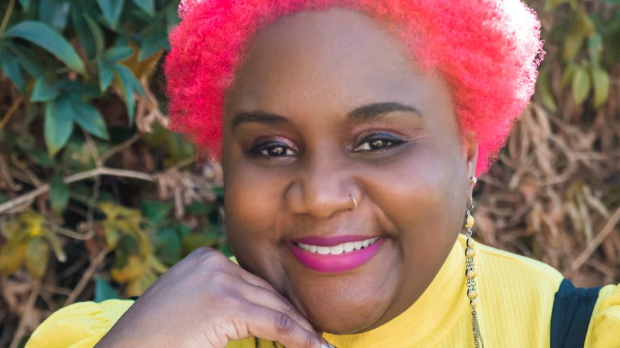 Nikki Bailey headshot smiling with pink hair wearing a yellow shirt