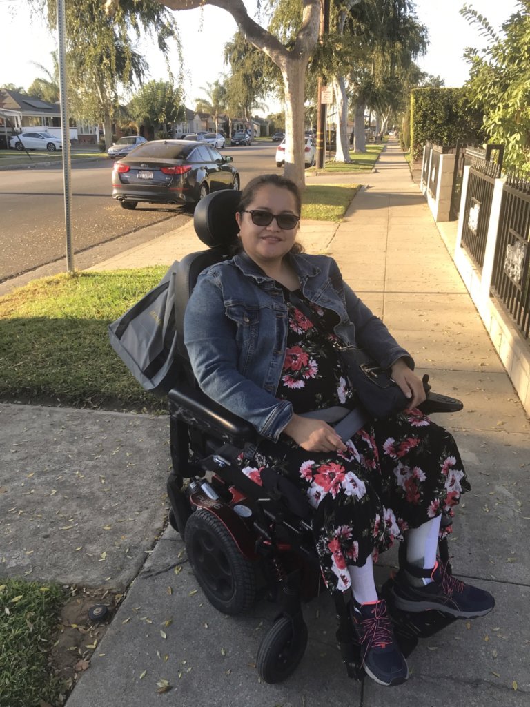 Nelly Nieblas smiling, seated in a wheelchair on a sidewalk
