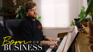 Chris playing piano in a scene from Born For Business. Show logo in bottom left.