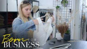 Lexi picking up a dress in a scene from Born For Business. Show logo in bottom left.