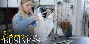 Lexi picking up a dress in a scene from Born For Business. Show logo in bottom left.