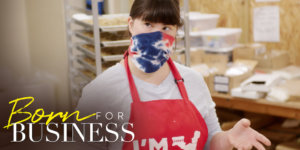 Collette Divitto in her bakery wearing a mask and apron in a scene from Born For Business. Show logo in bottom left