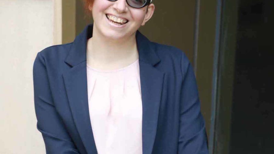 Lauren Arena smiling wearing glasses and a suit jacket. Arena is a wheelchair user.