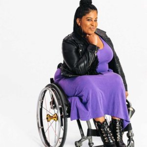 a brown skin black woman posing in her wheelchair with a purple dress and faux leather jacket. She is wearing her hair half up half down with long braids in a photo studio with a white backdrop