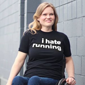Kirsten Sharp smiling seated in her wheelchair wearing a t-shirt that says "I hate running"