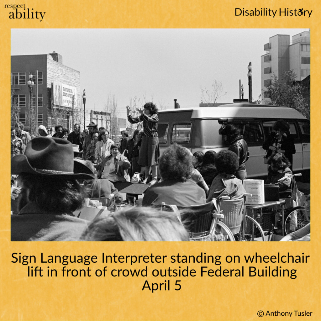 Black and white photo of crowd of protesters outside of the Federal Building. A sign language interpreter stands on a van wheelchair lift, signing to the crowd. Text: Sign Language Interpreter standing on wheelchair lift in front of crowd outside Federal Building. April 5. Source: Anthony Tusler. 