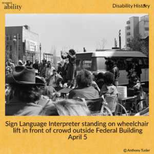 Black and white photo of crowd of protesters outside of the Federal Building. A sign language interpreter stands on a van wheelchair lift, signing to the crowd. Text: Sign Language Interpreter standing on wheelchair lift in front of crowd outside Federal Building. April 5. Source: Anthony Tusler.