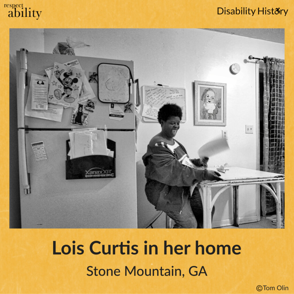 Black and white photo of Lois Curtis sitting at her kitchen table, smiling with a paper in her hand. To the left of her is her fridge and behind her is a white board and framed drawing. Text: Lois Curtis in her home. Stone Mountain, GA. Source: Tom Olin