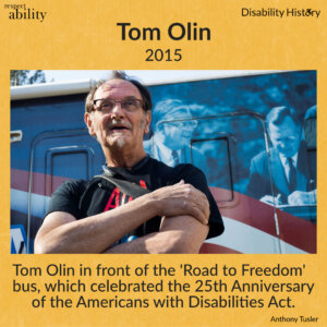 Color photo of Tom Olin standing with his arms crossed in front of the ‘Road to Freedom’ bus that has the American flag on it and Tom’s photo of George H.W. Bush signing the ADA. Text: Tom Olin in front of the Road to Freedom bus, which celebrated the 25th Anniversary of the Americans with Disabilities Act. Source: Anthony Tusler.