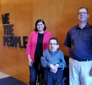 Steven Tingus, Lauren Appelbaum, and Delbert Whetter smile in front of a wall with text on it that says "We the People"