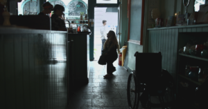 Still from "Is There Anybody Out There" with Ella Glendining standing next to her wheelchair in a dark store, speaking to people behind a desk.