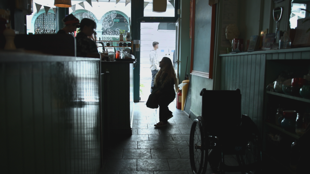 Still from "Is There Anybody Out There" with Ella Glendining standing next to her wheelchair in a dark store, speaking to people behind a desk.