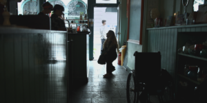 Still from "Is There Anybody Out There" with Ella Glendining standing next to her wheelchair in a dark store, speaking to people behind a desk.