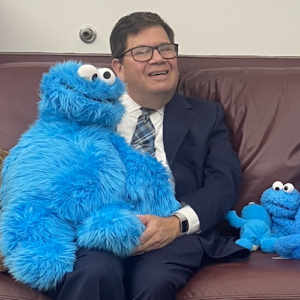 Rabbi Alan Cook holding a cookie monster puppet seated on his couch, smiling