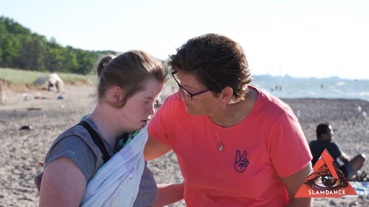 A scene from "Teacher of Patience" with Emily and a woman talking on a beach. Slamdance logo in bottom right corner.