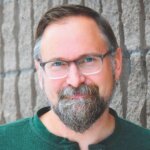 Keith Dow headshot wearing glasses and a green shirt in front of a stone wall