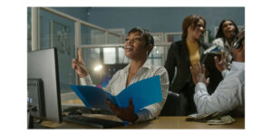 Still from "Oreo" featuring a black woman holding a folder sitting in an office as co-workers are talking behind her