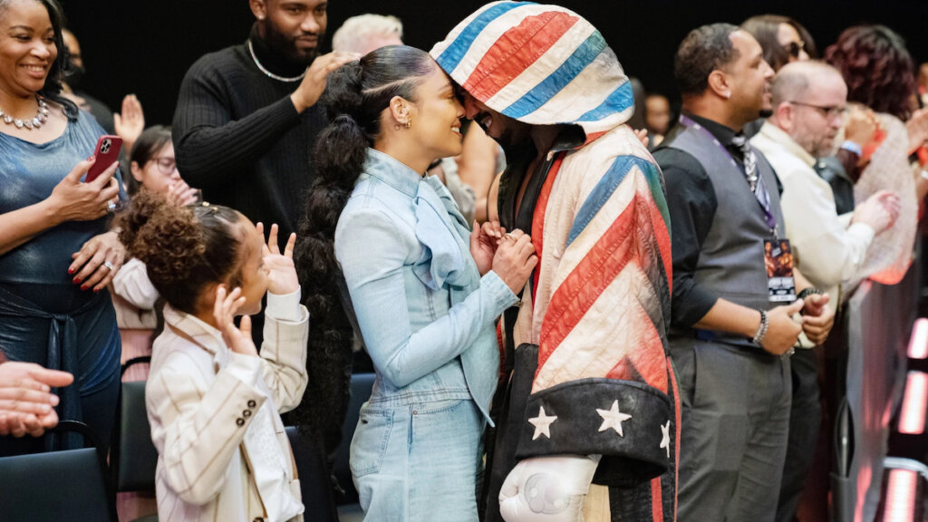 a still from CREED III with Mila Davis-Kent cheering for Michael B. Jordan after a fight as he kisses Tessa Thompson.