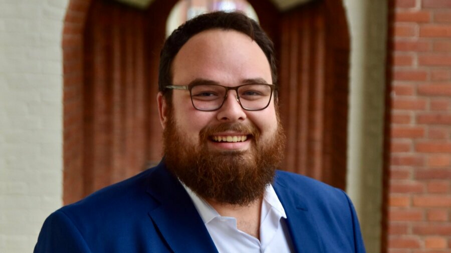 Ben Bond smiling headshot in front of a blurred background wearing a blue suit jacket.