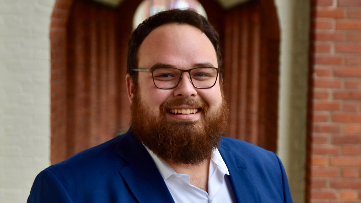 Ben Bond smiling headshot in front of a blurred background wearing a blue suit jacket.