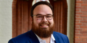 Ben Bond smiling headshot in front of a blurred background wearing a blue suit jacket.