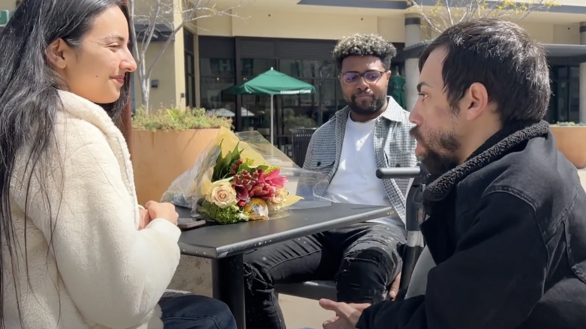 still from "Unexpected Date" featuring Nader Bahu, Dante Fontenot, and Vikki Beretta around a table outside after Bahu just gave Beretta flowers