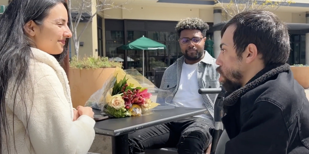 still from "Unexpected Date" featuring Nader Bahu, Dante Fontenot, and Vikki Beretta around a table outside after Bahu just gave Beretta flowers