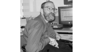 Neil Jacobson sitting at his computer and smiling he has a beard and is wearing glasses grayscale photo