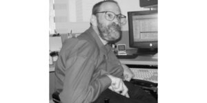 Neil Jacobson sitting at his computer and smiling he has a beard and is wearing glasses grayscale photo