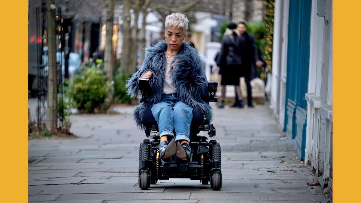 Still from "My Eyes Are Up Here" with a person using a power wheelchair to navigate down a city sidewalk