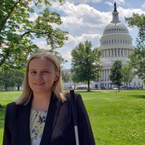 Stacy Cervenka smiling wearing a suit and holding her white cane with the U.S. Capitol building in the distance behind her