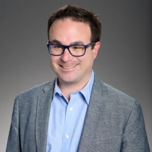 Nathan Varni smiling headshot wearing a blue shirt and grey blazer