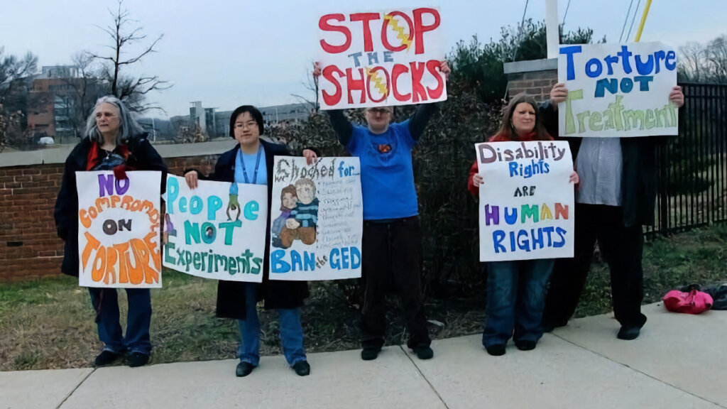 Protesters holding signs that say things like "stop the shocks," "disability rights are human rights," and "people not experiments"