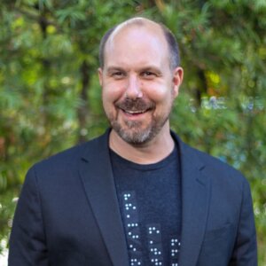 Chad Allen smiling wearing a blazer and a t-shirt with braille on it