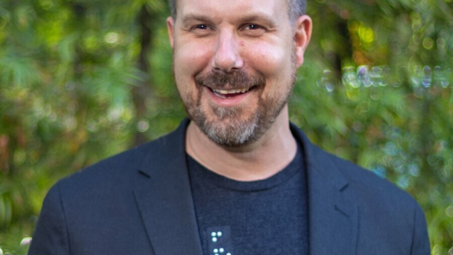 Chad Allen smiling wearing a blazer and a t-shirt with braille on it