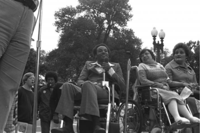 black and white photo of Judy Heumann - a white woman sitting in her wheelchair - at the 504 sit in. Next to her on the left is a member of the Black Panther Group Bradley Lomax. To her right is another disability rights activist.