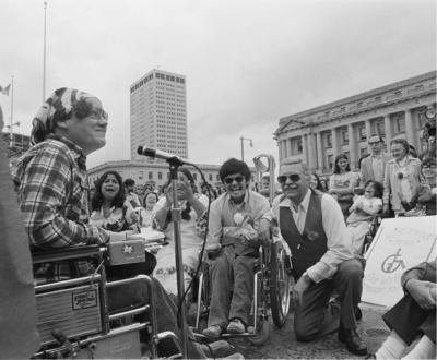 Kitty Cone, a disability rights activist, seated in her wheelchair with a crowd of other activists listening to her