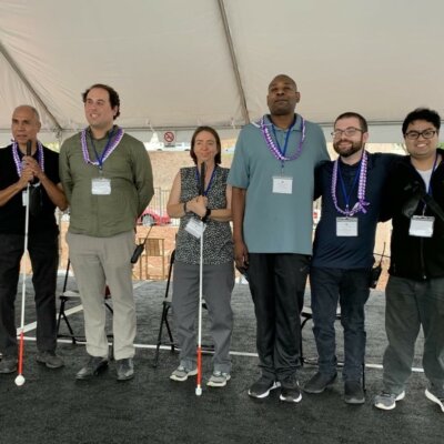 Six former RespectAbility Fellows stand on stage under a tent holding their white canes