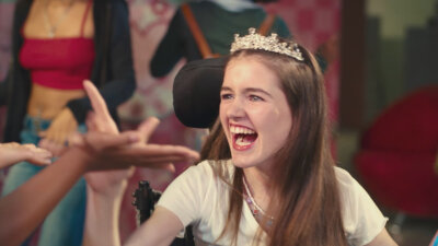 Phoebe-Rae Taylor smiling wearing a crown seated in her wheelchair in a scene from Out of My Mind