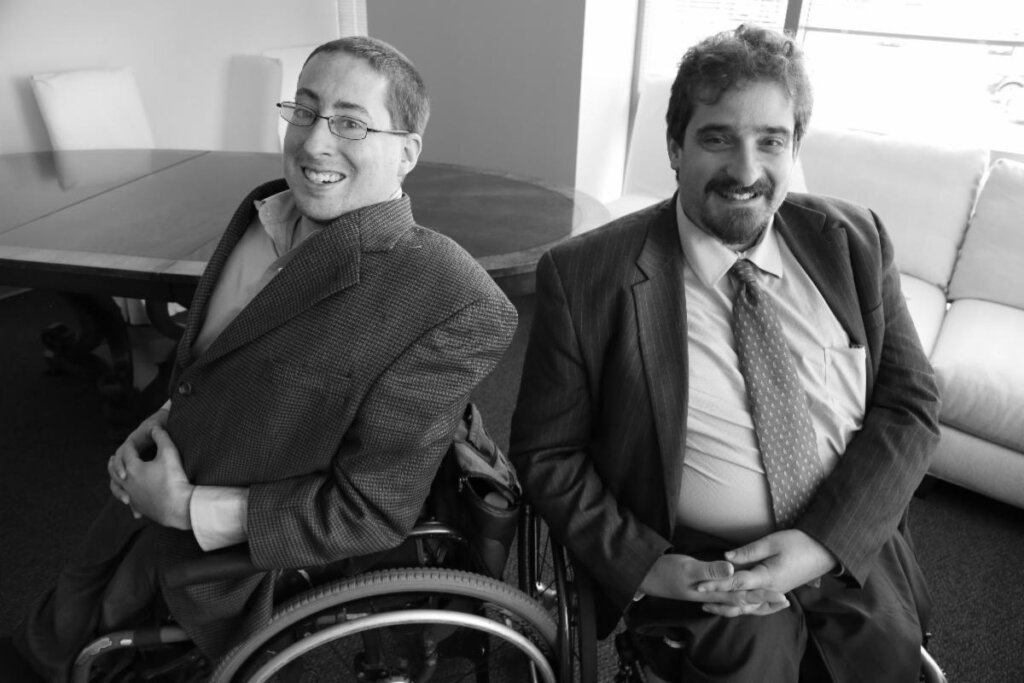 Ben Spangenberg and Justin Chappell smiling wearing suits seated in their wheelchairs in an office