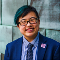 Lydia X.Z. Brown smiling headshot wearing a blue suit, glasses, and a blue patterned tie