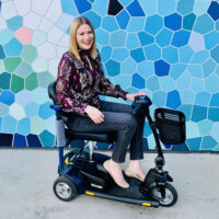 Amy, a disabled, white Australian woman with blonde hair, sits on her mobility scooter in front of a wall with blue and white geometric shapes. 
